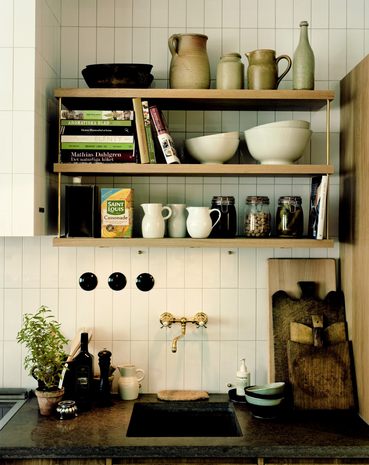 Ett Hem hotel kitchen with open wooden shelving, ceramic vessels, books, and brass fixtures above a dark countertop.