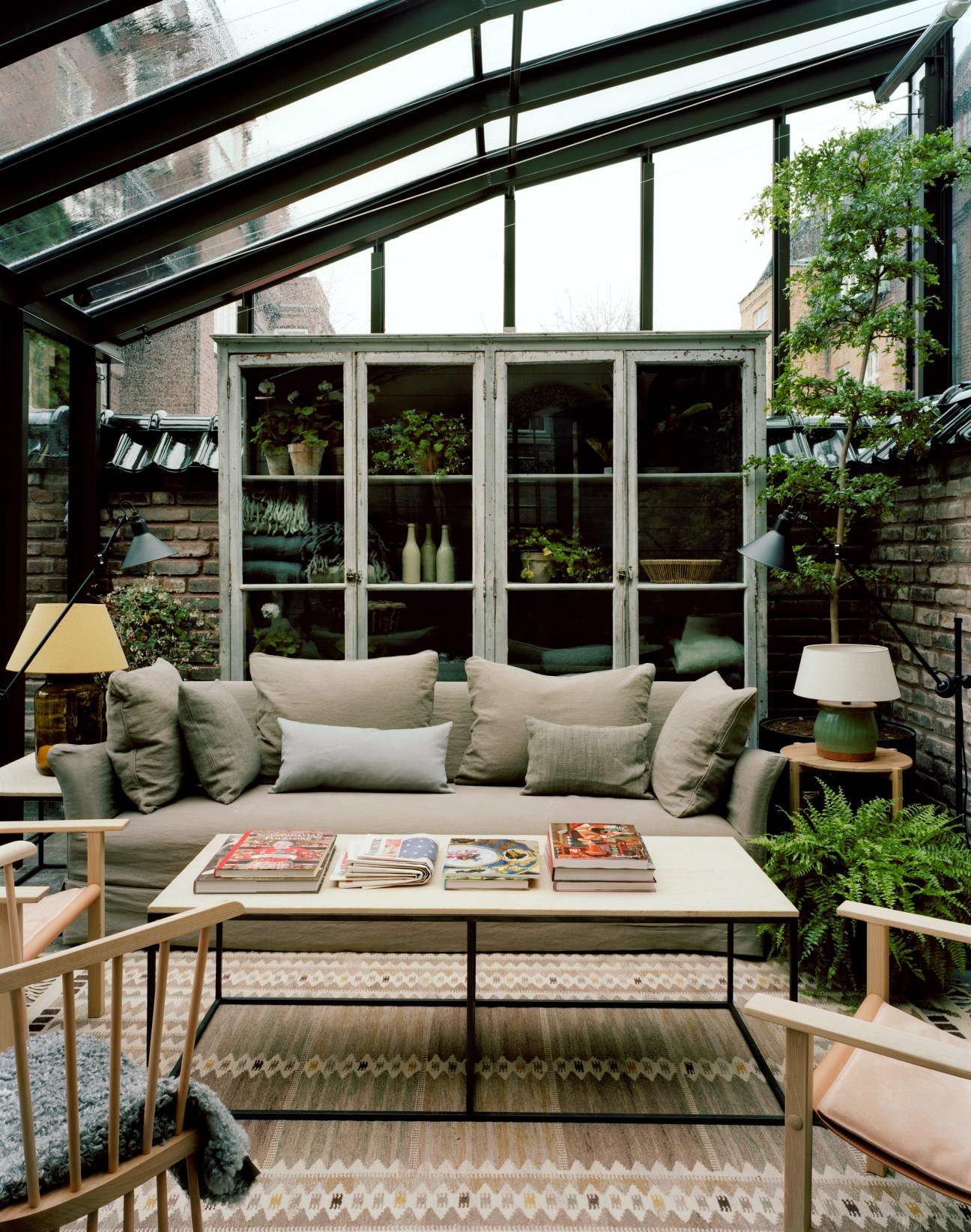 Glass conservatory with green-framed doors, comfortable seating, plants, and magazines on a table.
