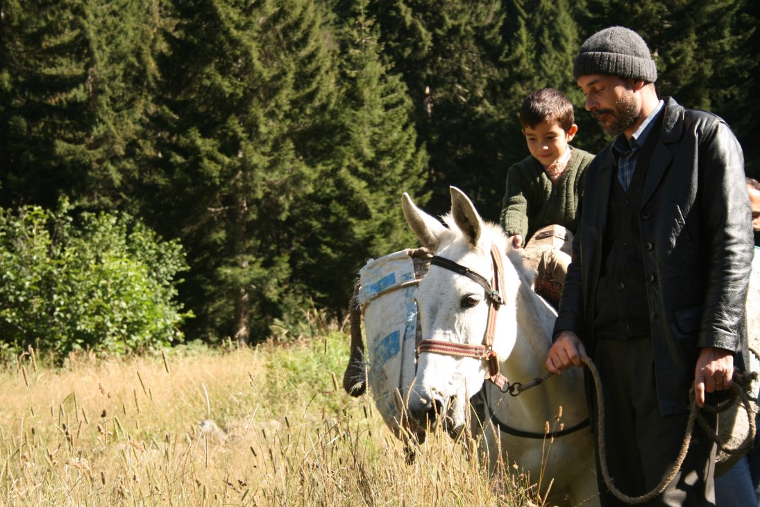 A man and young boy stand with a white horse in a forested mountain landscape.