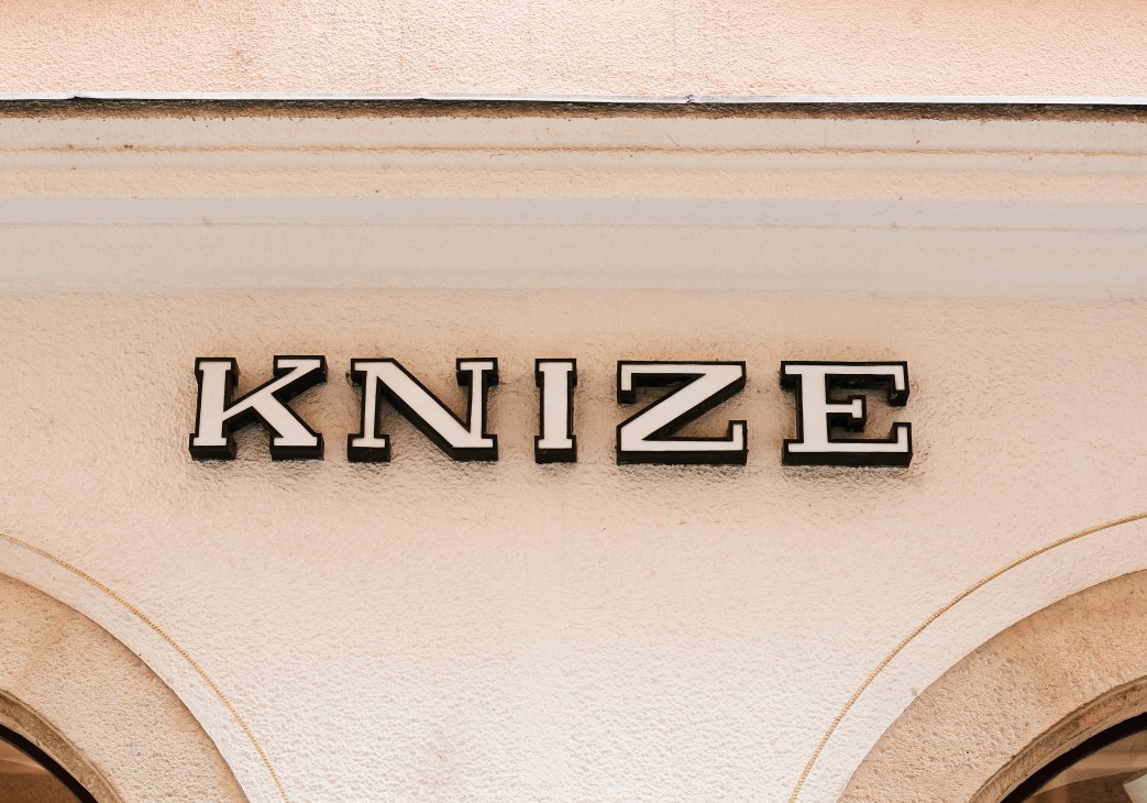 Vintage "KNIZE" storefront sign in outlined lettering on a cream-colored façade in Vienna.