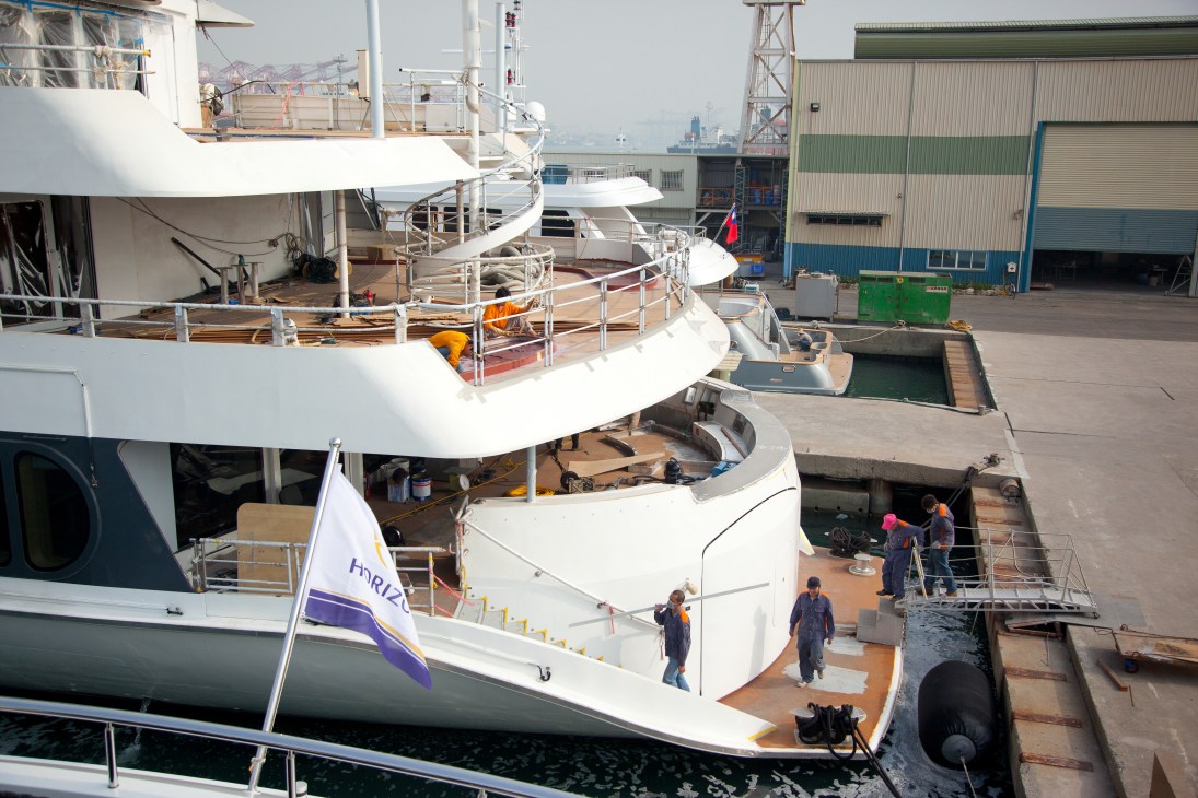 Luxury yacht under construction at Horizon shipyard in Kaohsiung with workers on deck and industrial buildings in background.