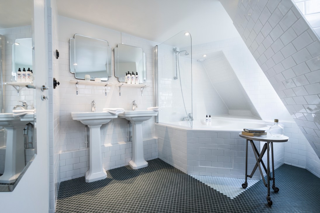 Modern bathroom with dual pedestal sinks, mirrors, white subway tile walls, and freestanding bathtub under sloped ceiling.