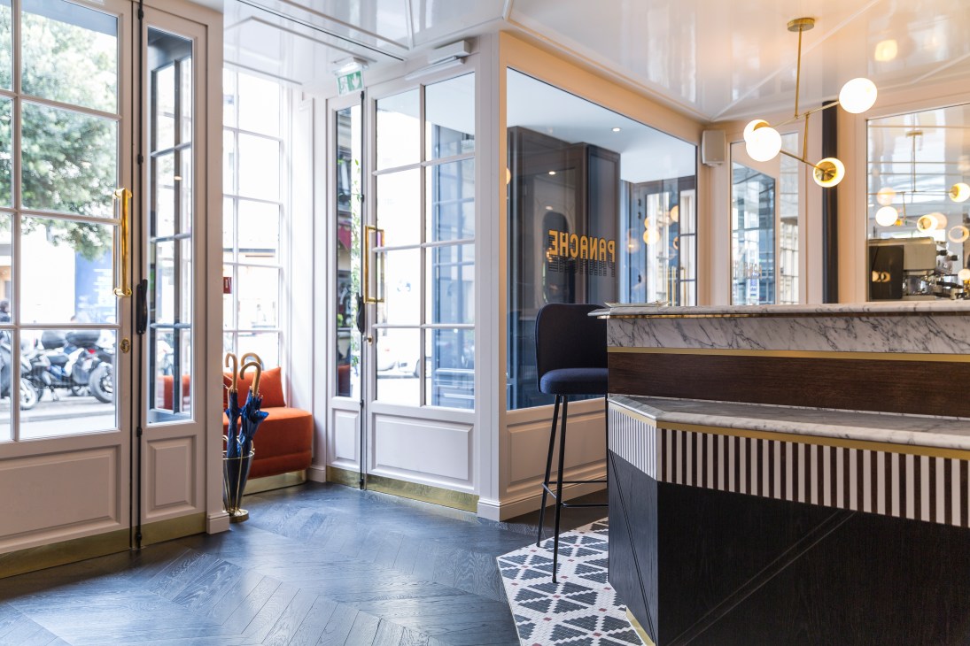 Hotel Panache Paris reception area with marble counters, brass fixtures, pendant lights, and street-facing windows.
