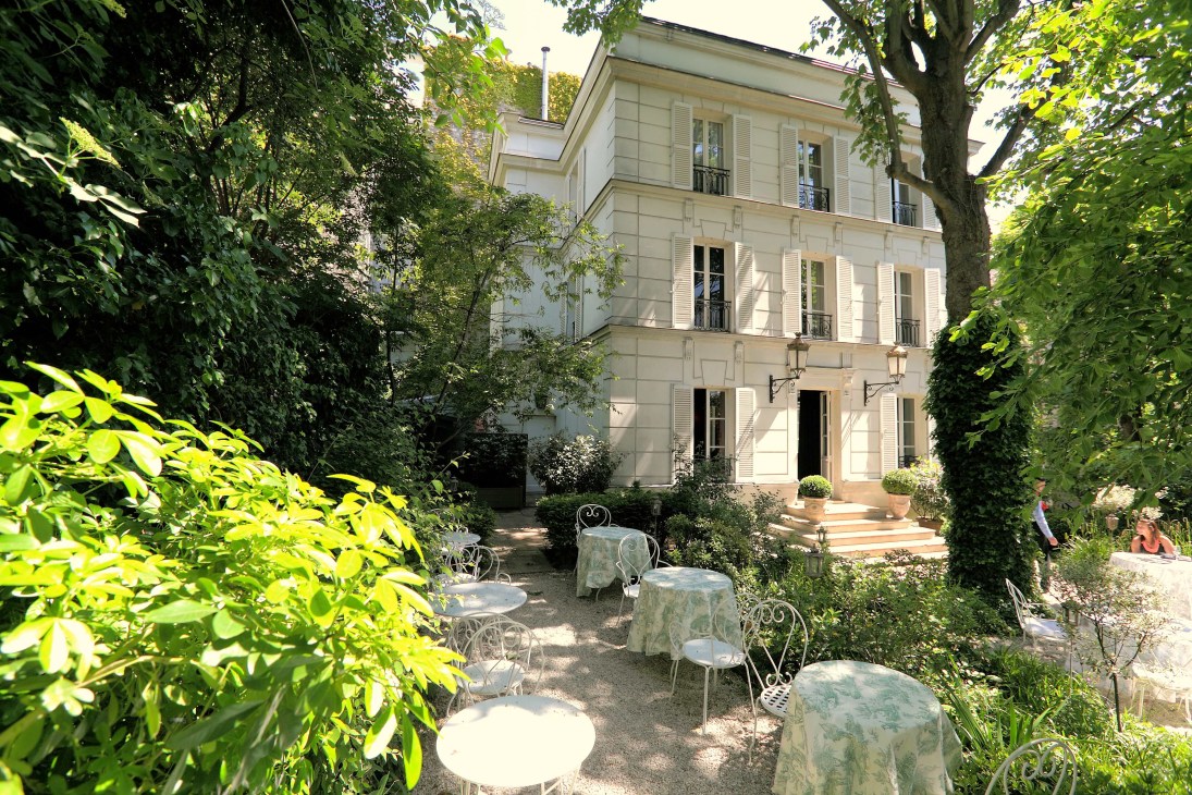 Cream-coloured mansion facade surrounded by trees with garden seating area featuring round tables and wrought iron chairs.