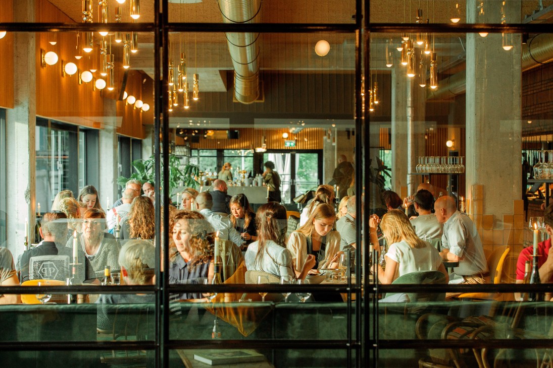 Diners enjoy drinks and conversation at Hotel V Fizeaustraat's restaurant, visible through large glass windows with hanging pendant lights.