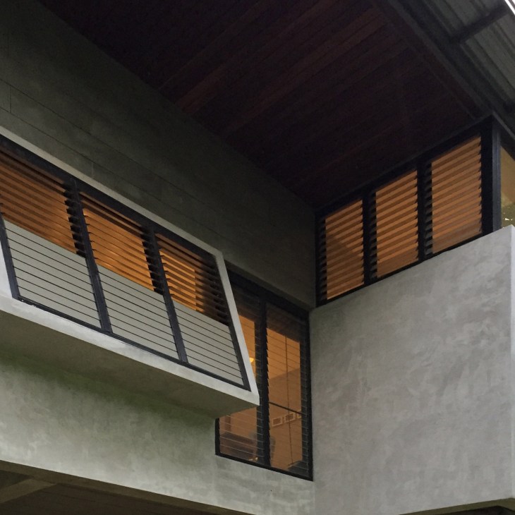 Wooden louvered windows with black frames on a concrete wall under a sloped roof.