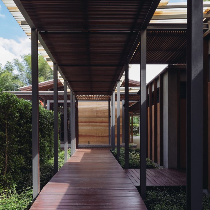 Covered wooden walkway with black structural beams and green foliage, leading to a wooden entrance door.