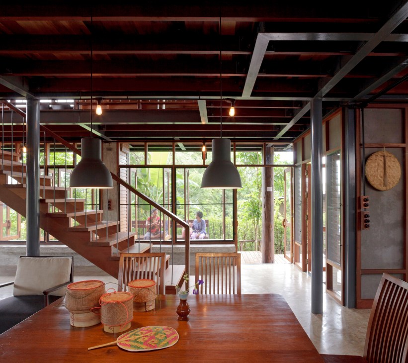 Interior of Udonthani house with wooden dining table, spiral staircase, large windows overlooking jungle, and pendant lights.