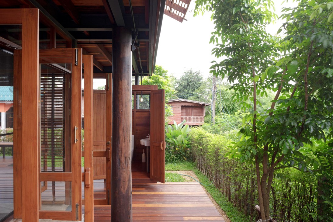 Timber-built Thai house on stilts with wooden deck and lush jungle vegetation surrounding it.