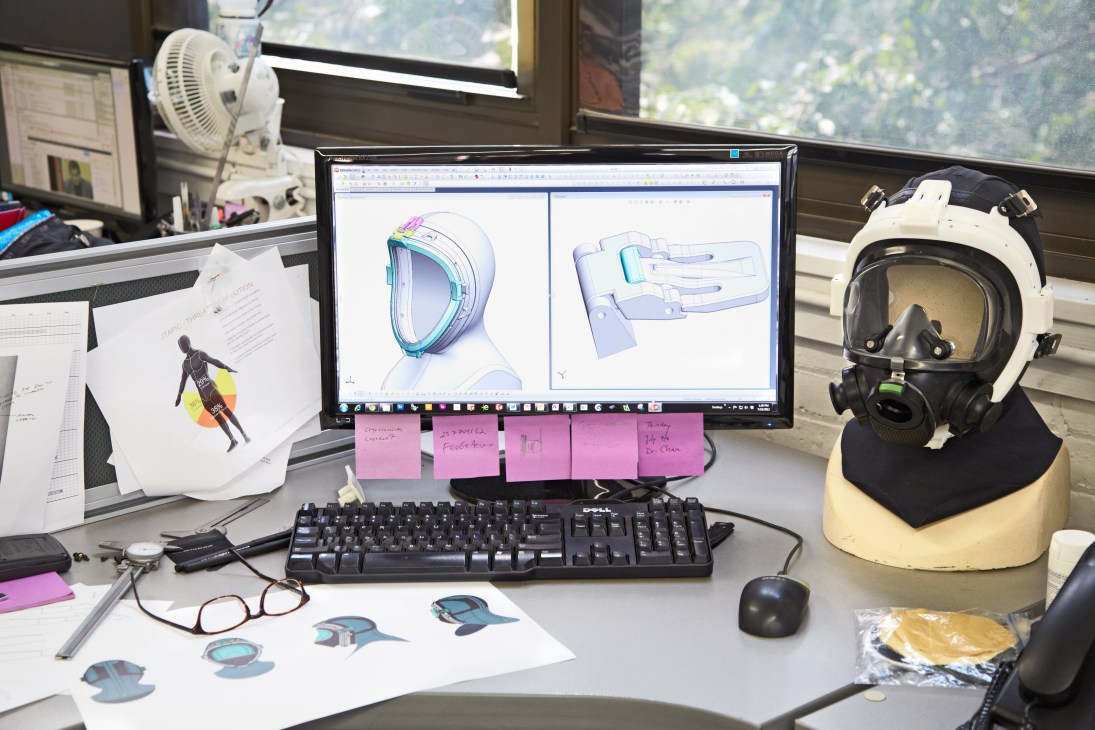 Designer's desk with computer displaying CAD helmet designs, sketches, prototypes, and pink sticky notes.