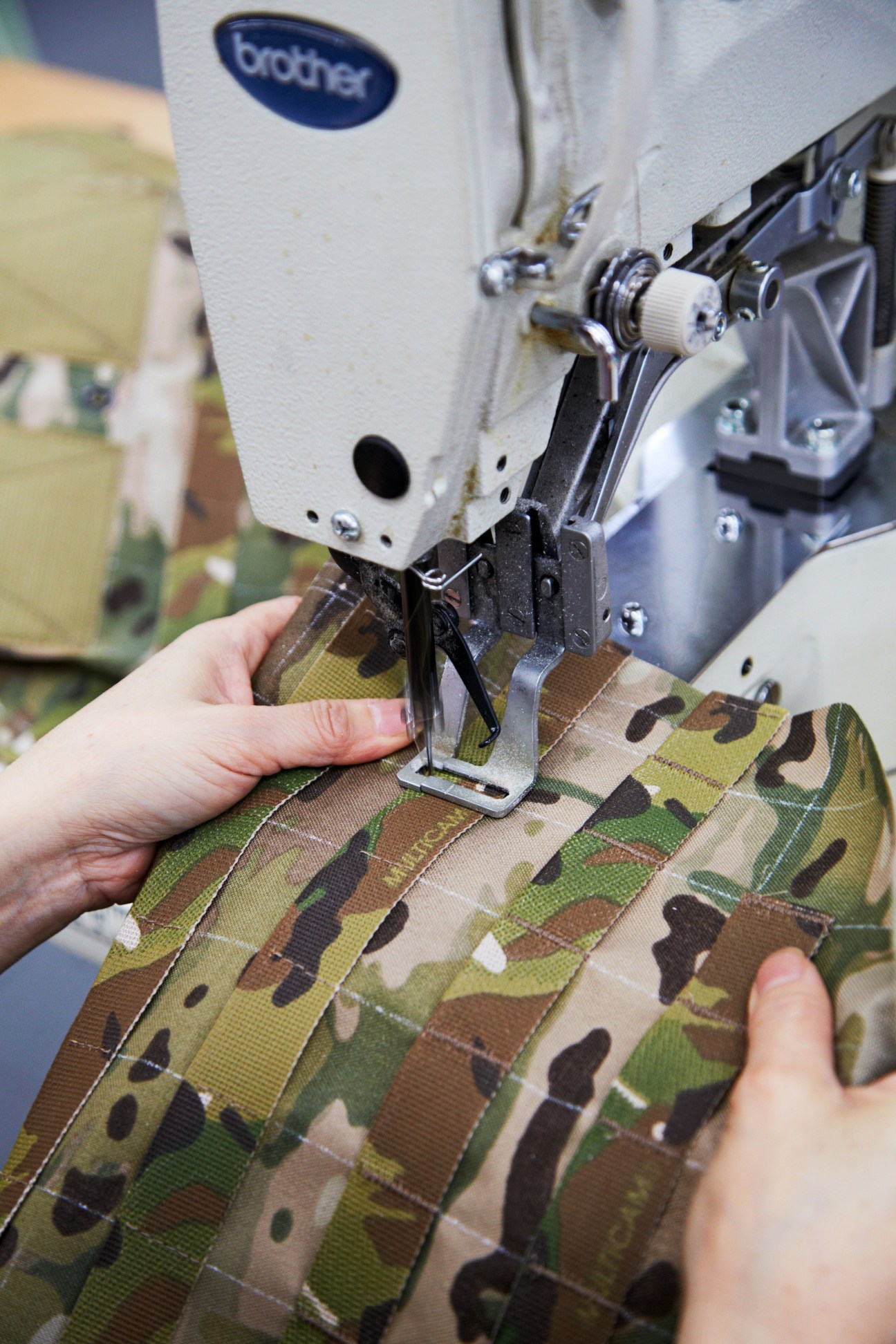 Worker sewing MultiCam patterned webbing on a Brother industrial sewing machine.