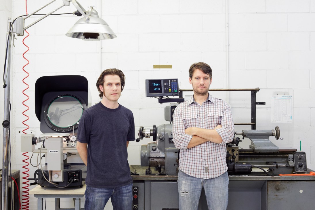 Caleb Crye and Gregg Thompson stand in their Brooklyn workshop surrounded by precision manufacturing equipment.