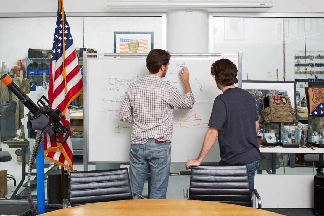 Two men design military equipment at a whiteboard in Crye Precision's Brooklyn office, surrounded by camouflage samples and military gear.