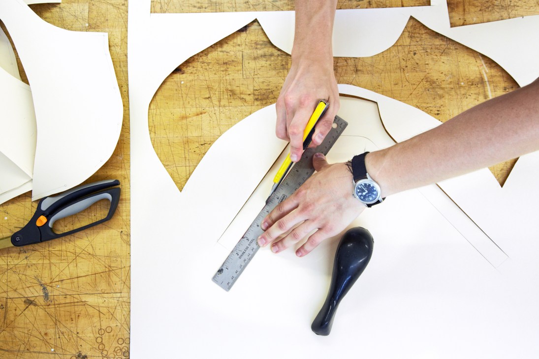 Hands measuring and marking white fabric with a ruler and pencil at a wooden worktable.