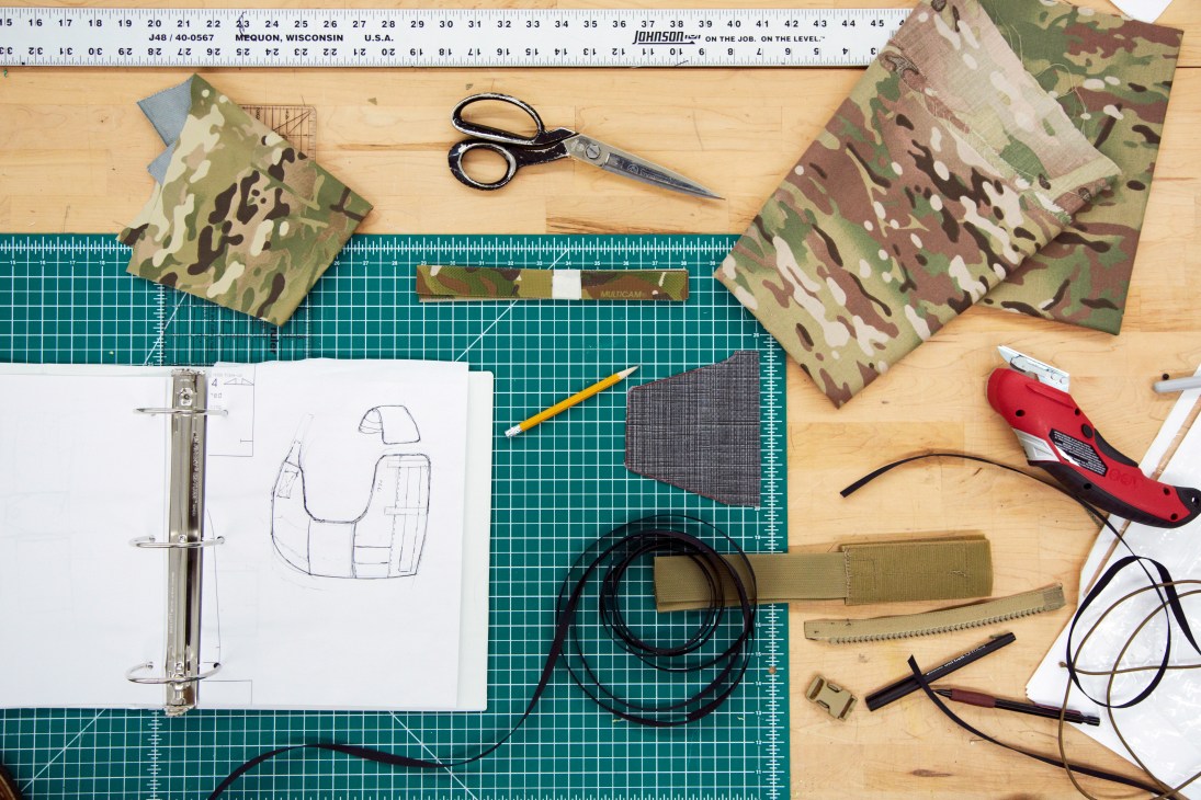 Design workspace with MultiCam fabric swatches, sketches, scissors, and prototyping tools on cutting mat.
