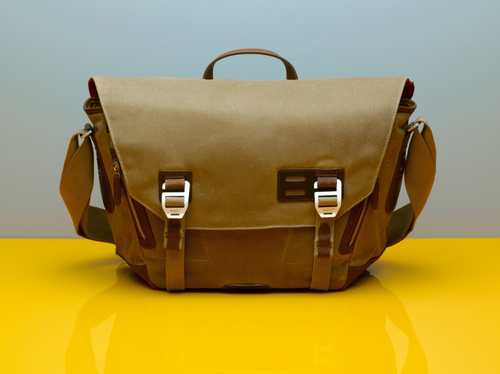 Howies tan canvas and leather messenger bag with buckle straps against blue and yellow background.