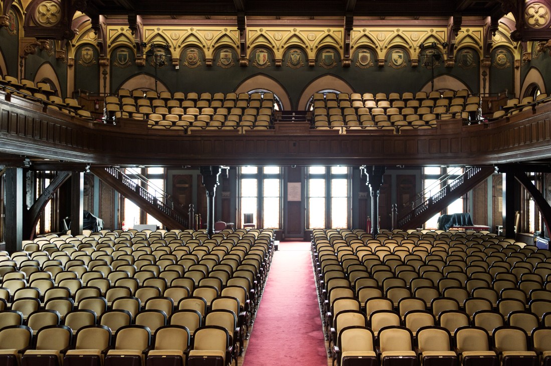Gaston Hall auditorium with ornate gold ceiling, rows of tan seats, and red carpet center aisle.