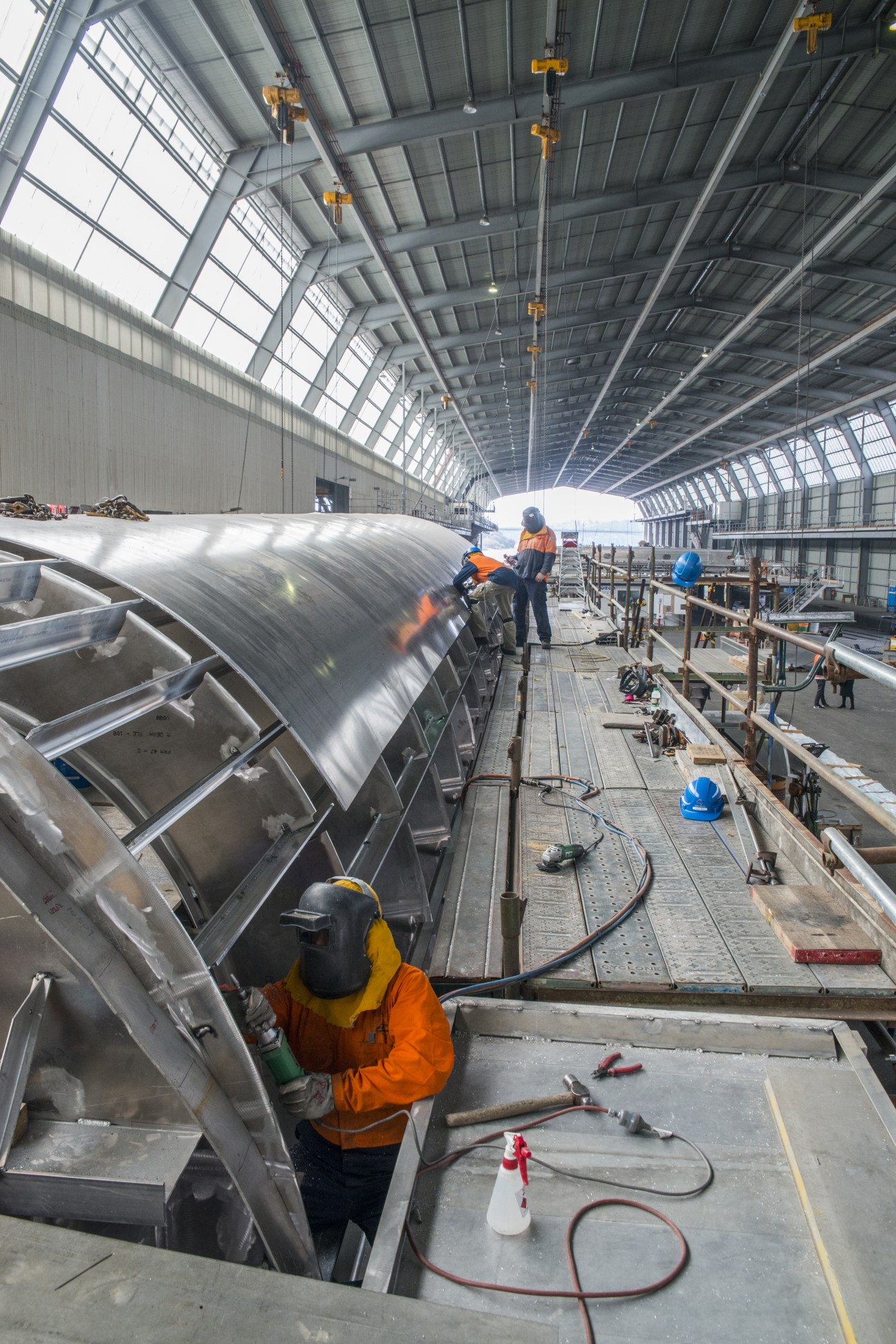 Welder in orange safety gear working on a large curved aluminium hull inside Incat's industrial shipyard shed.