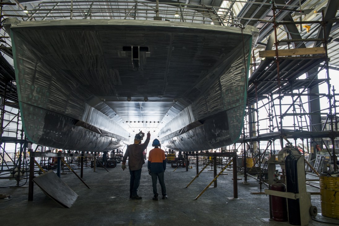 Two workers inspect a large aluminium catamaran hull under construction inside Incat's shipyard.