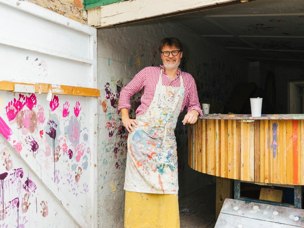 Simon March stands in his paint shop wearing a paint-splattered apron and checkered shirt, smiling at the camera with colorful paint swatches visible on the walls behind him.