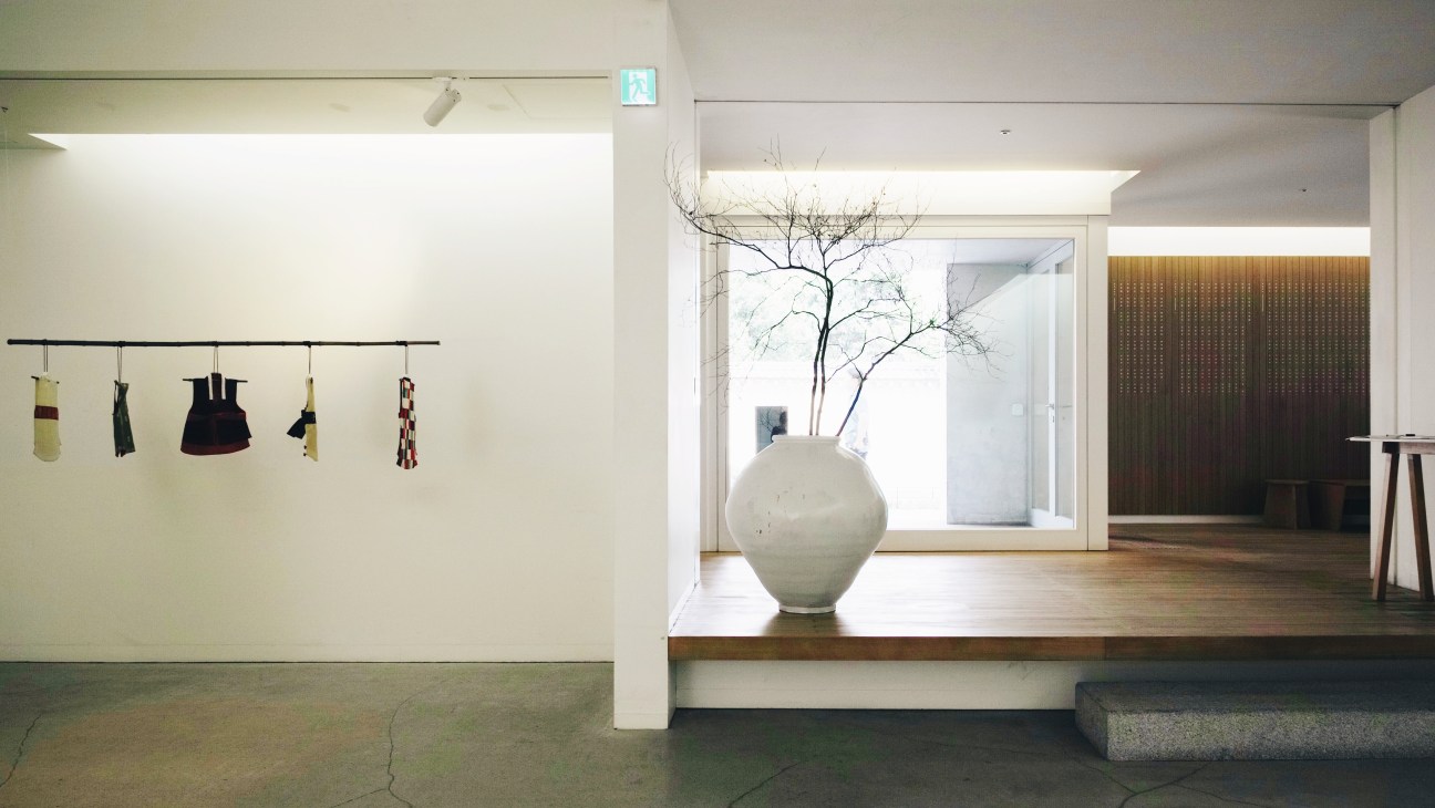 Minimalist exhibition space with traditional Korean clothing hanging on wall and large ceramic vessel displayed near window.