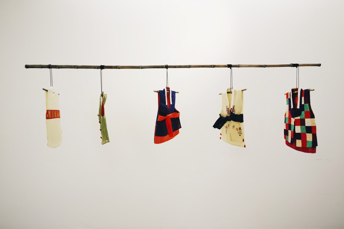 Traditional children's costumes by Lee Hong Soon hanging from a bamboo rod against a white wall.
