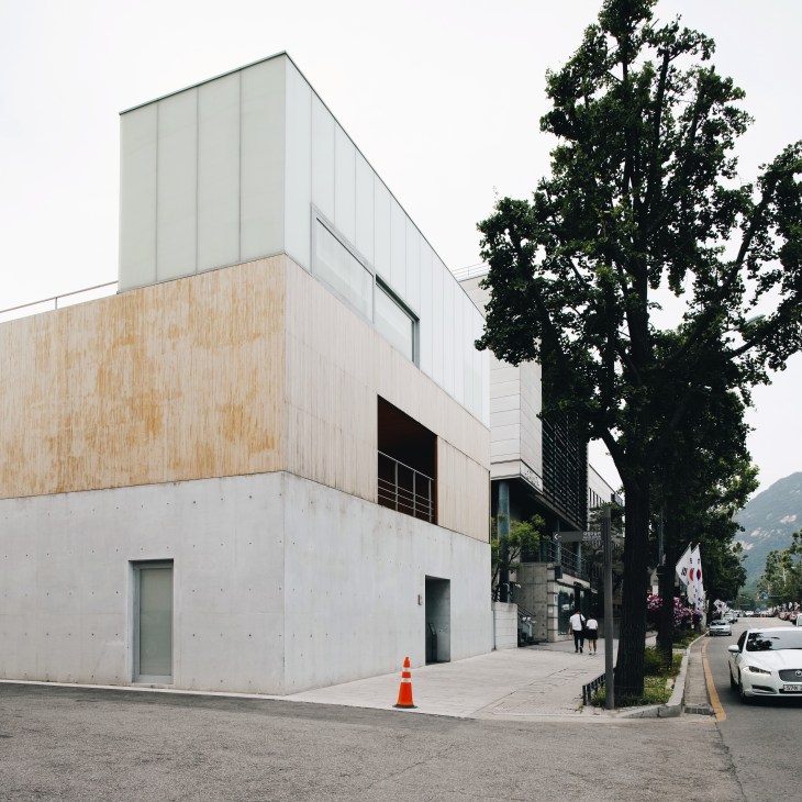Modern multi-level building with white and wood paneling exterior, located in Seoul near a large tree.