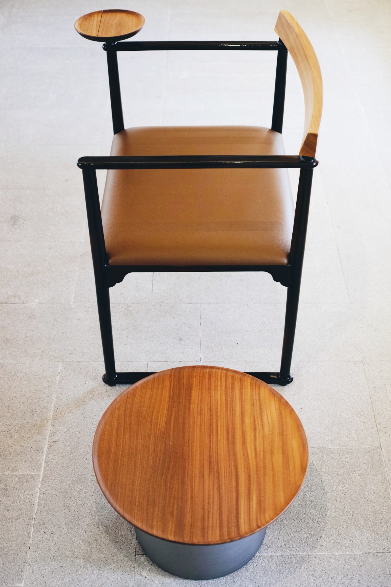 Minimalist chair with black frame, tan leather seat, and wooden armrest and backrest, displayed with a round wooden table below.