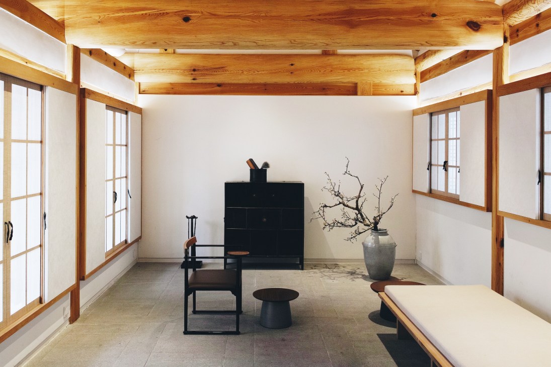Interior of a reconstructed traditional Korean hanok home with wooden beams, paper windows, and minimalist furniture including black chairs and a ceramic vessel.