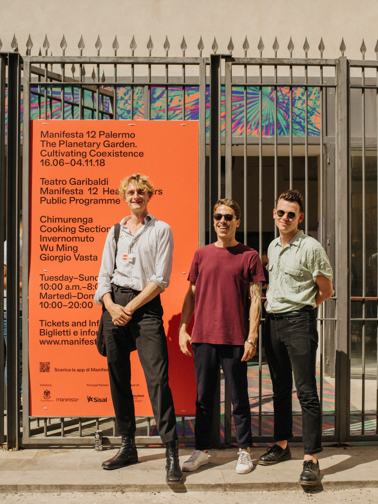 Three men standing in front of a metal gate with a large orange Manifesta 12 Palermo poster and colorful artwork visible behind them.