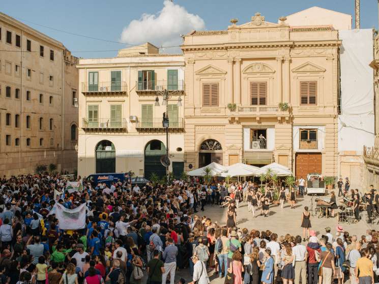Large crowd of people walking through a historic Palermo square lined with neoclassical buildings during the Palermo Procession event.