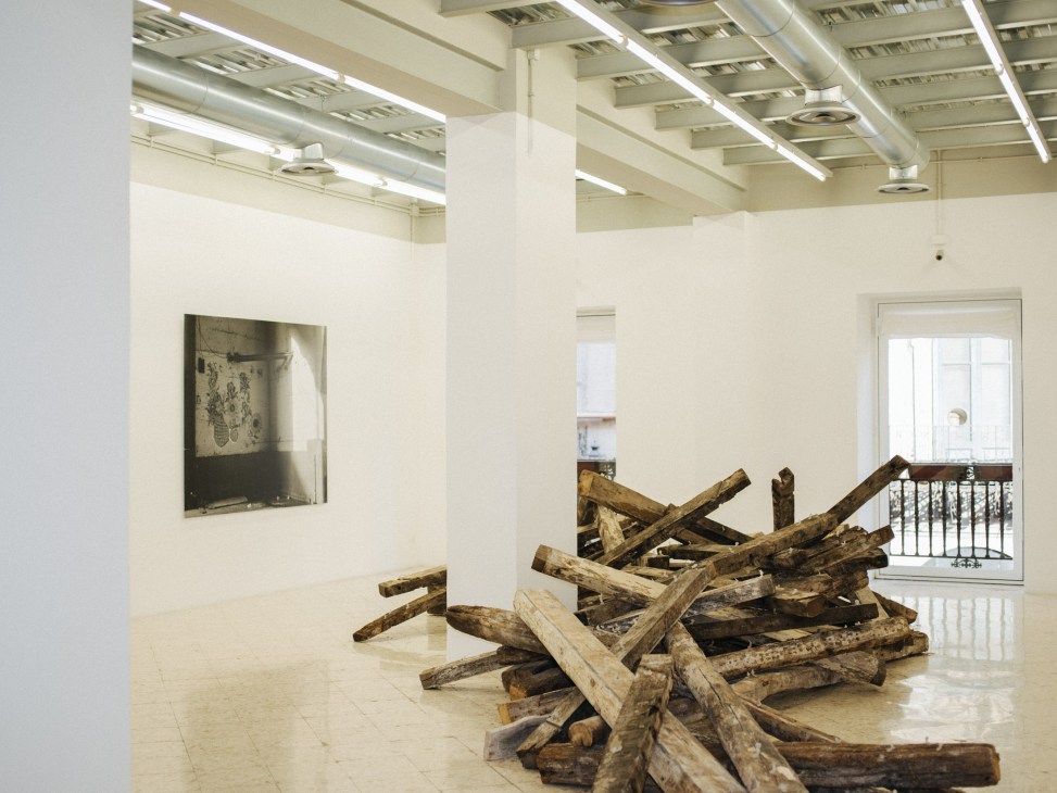 Contemporary art installation with wooden logs arranged in a minimalist gallery space with white walls and industrial ceiling