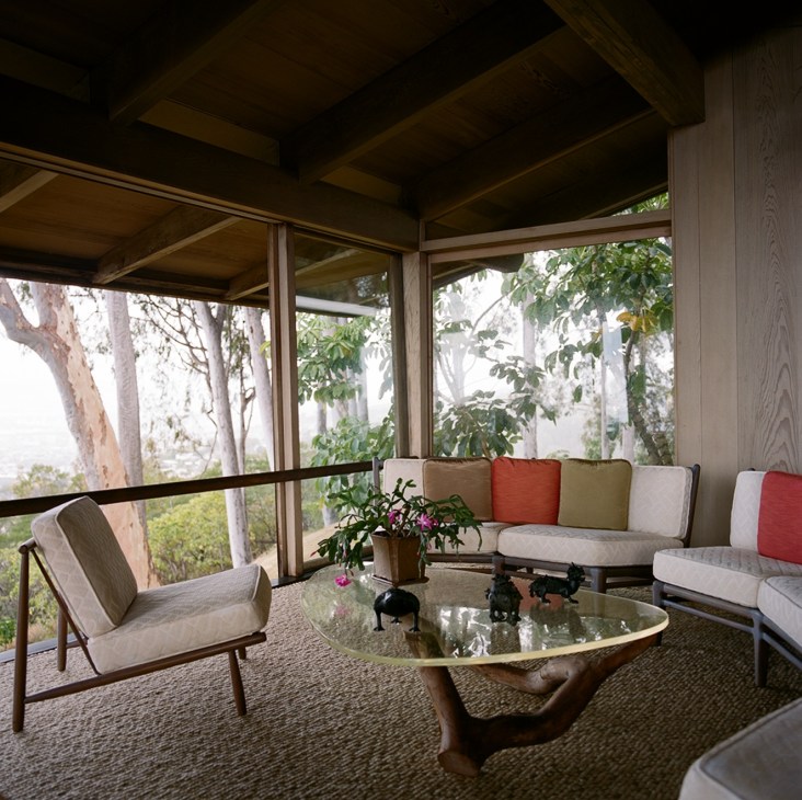 Mid-century modern living room with floor-to-ceiling windows overlooking trees, featuring a sculptural coffee table and colorful cushioned seating.