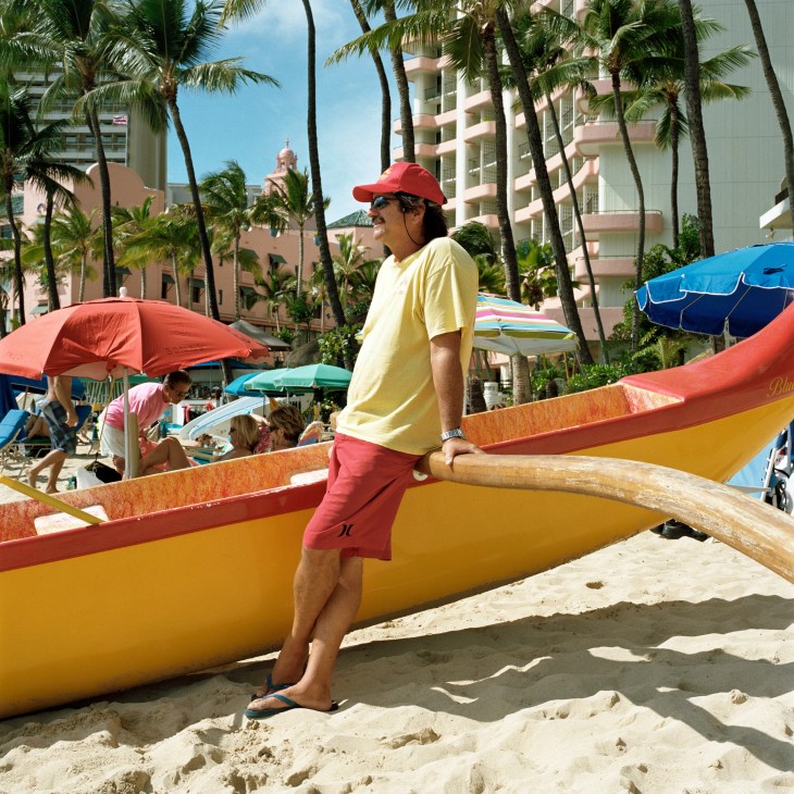 Man in yellow shirt and red cap stands with outrigger canoe on Waikiki beach with hotels and umbrellas in background.