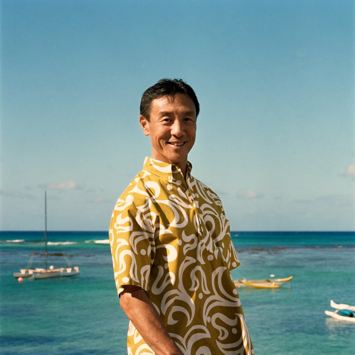 Michael Ako, general manager of Outrigger Canoe Club, smiles at the camera wearing a yellow and white patterned Hawaiian shirt with the ocean and outrigger canoes behind him.