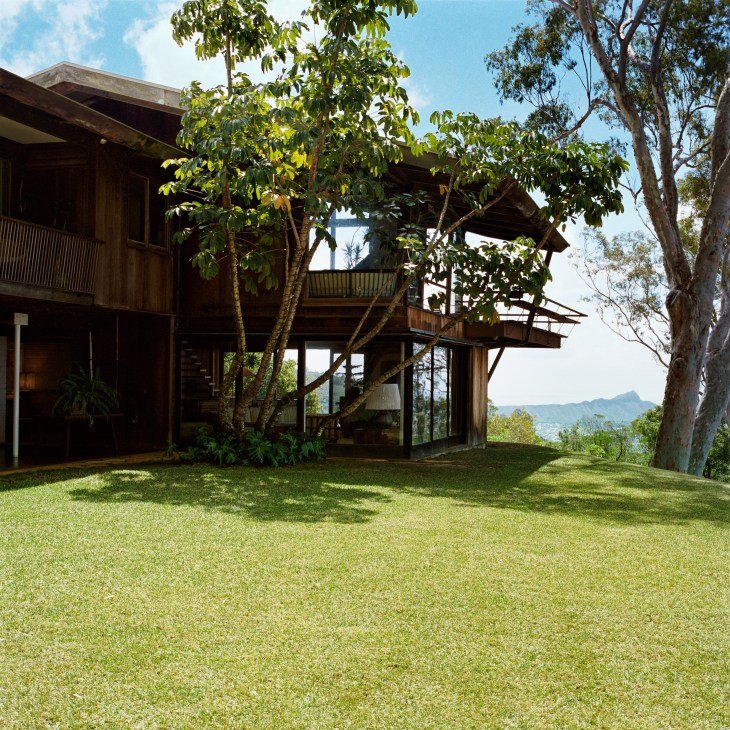 Modernist wooden house with open decks nestled among tropical trees and manicured lawn overlooking mountains.