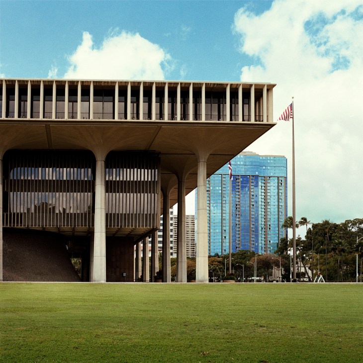 Brutalist concrete government building with columns and upper-level colonnade, American flag visible, modern high-rises in background.