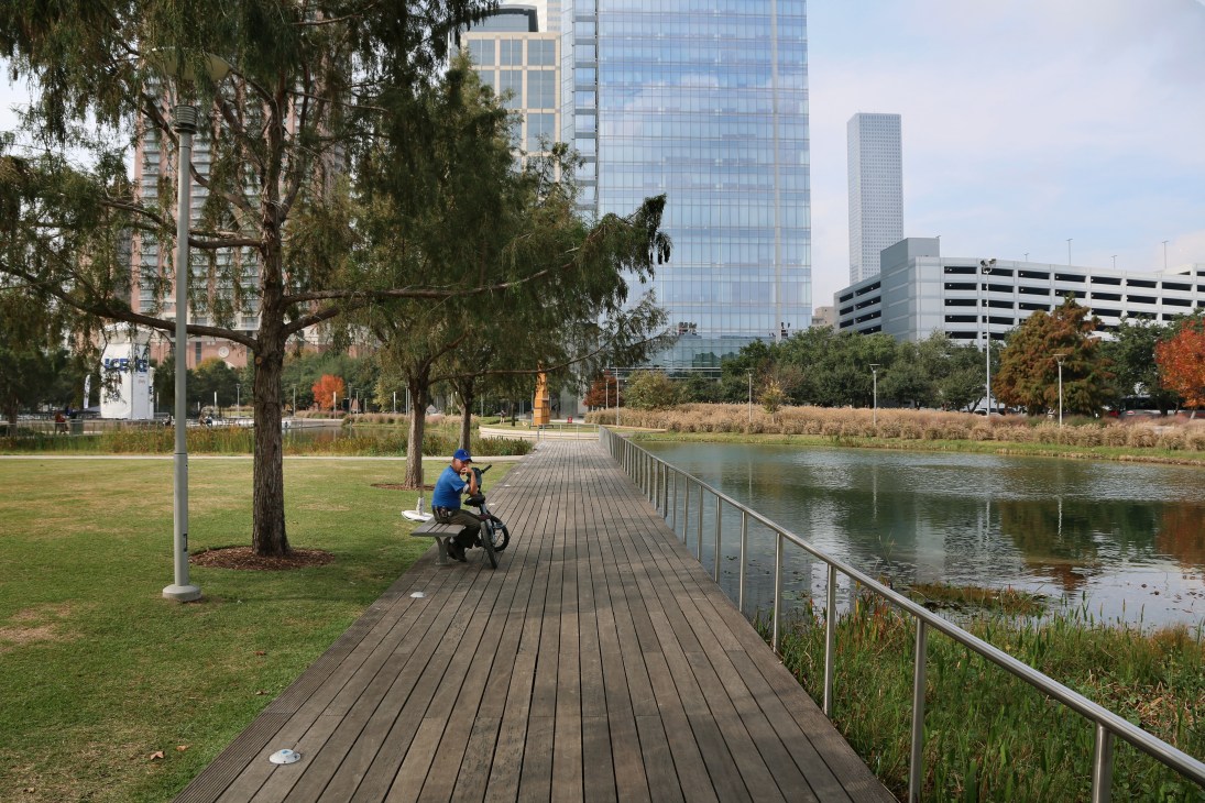 Person on bicycle riding along wooden boardwalk at Discovery Green park with water, trees and Houston skyscrapers in background.