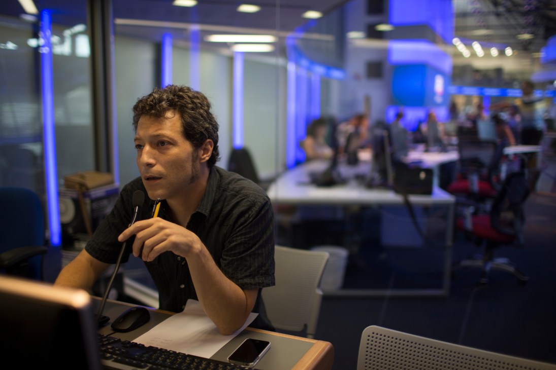 Male journalist at i24 News newsdesk holding pen, with blue-lit open newsroom visible behind him.