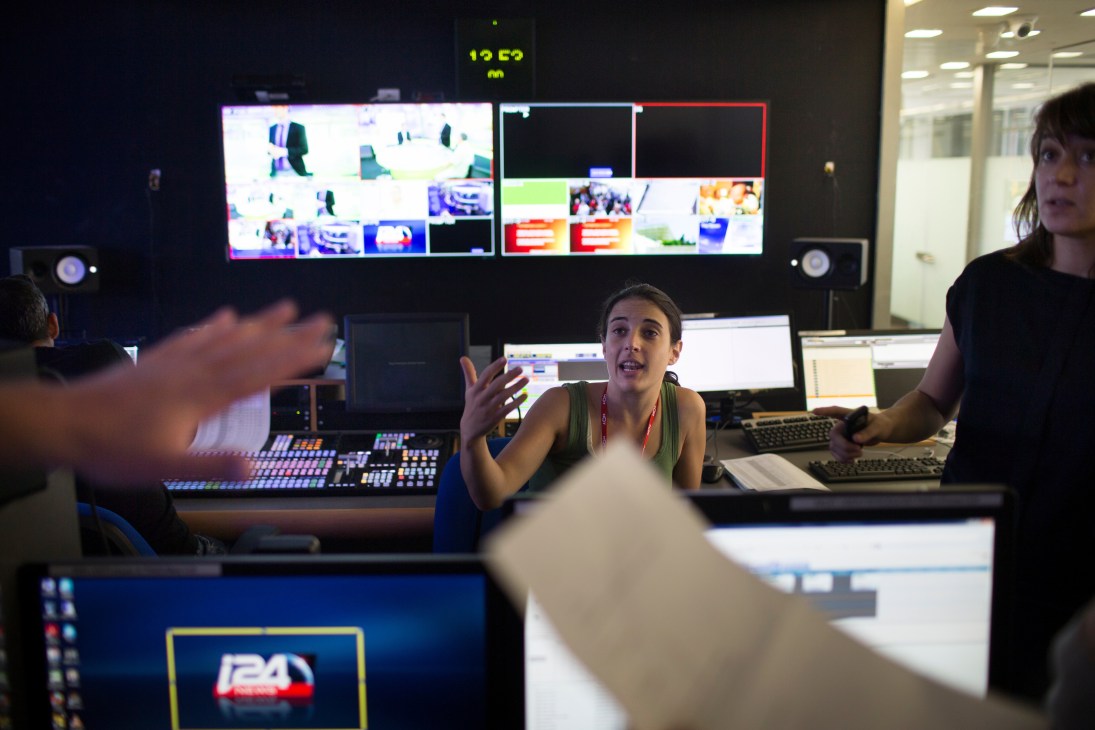 Staff members in i24 News control room with multiple broadcast monitors displaying video feeds and graphics.
