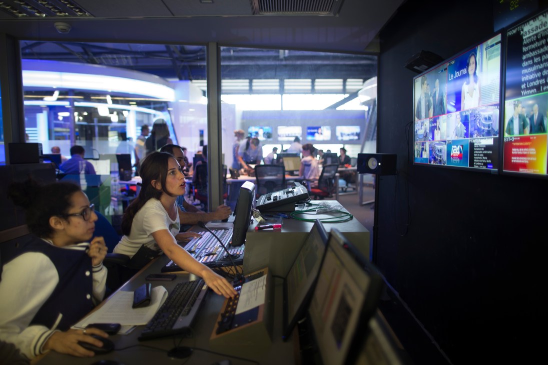 Staff members work at computer terminals in i24 News control room with multiple monitors displaying news feeds.