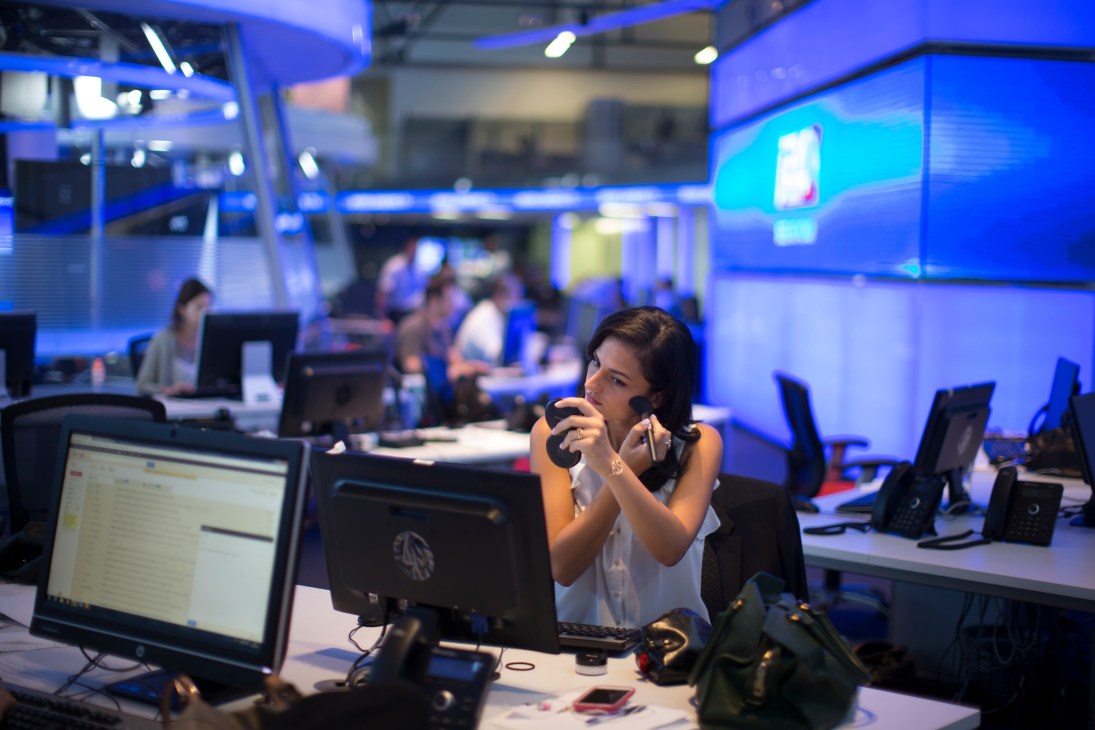 A woman in a white sleeveless top applies makeup at a desk in a modern newsroom with blue lighting and multiple computer monitors.