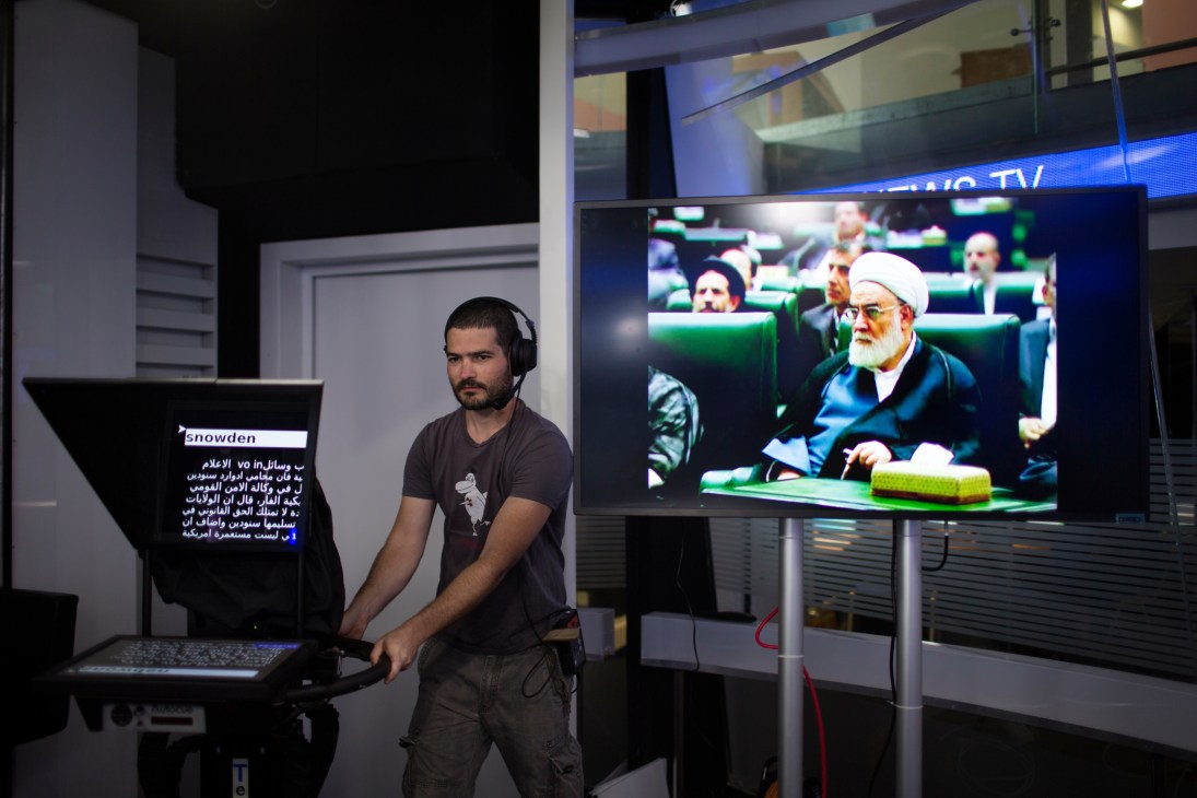 Journalist wearing headphones operates equipment at i24 News studio with monitor displaying broadcast content.