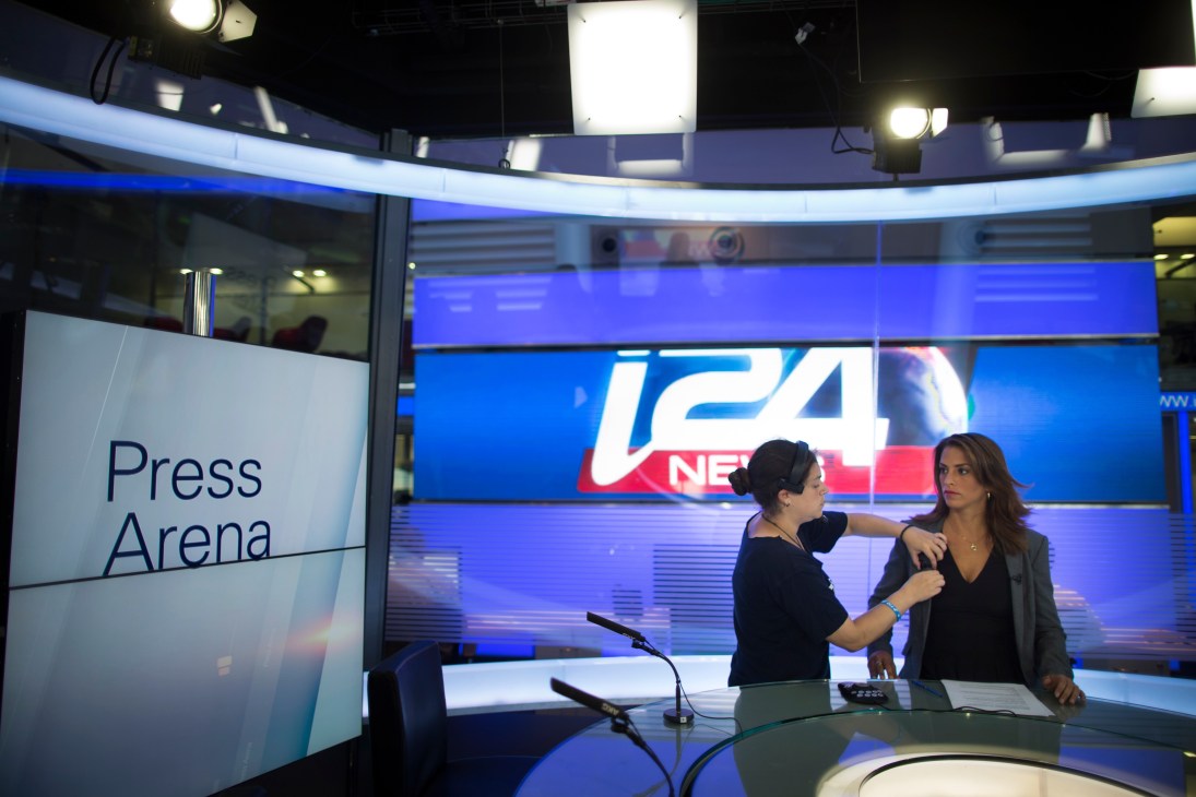 News anchor Hadas Sinai prepares at an i24 News studio desk with blue branding visible behind her.
