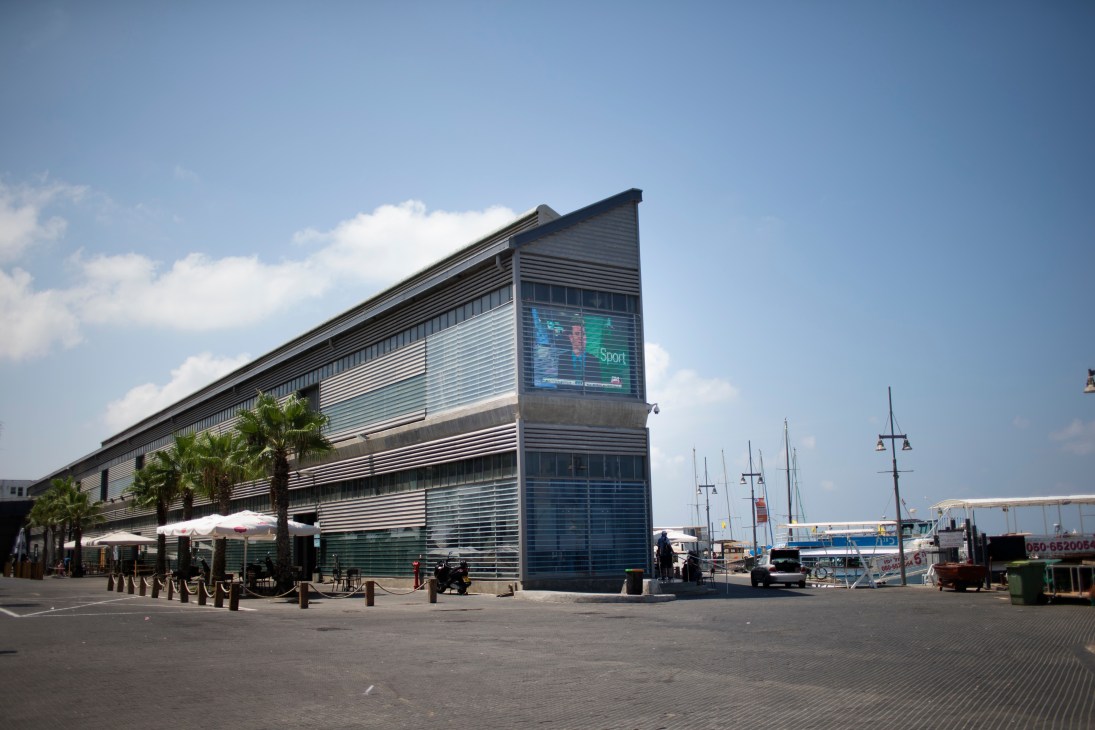 Modern glass and metal building with horizontal slats housing i24 News headquarters at Jaffa port with boats visible.