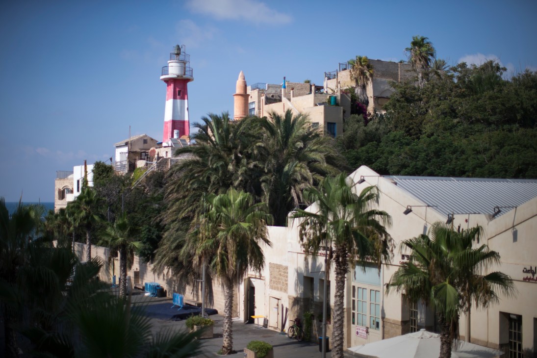 Street scene in Jaffa with palm trees, historic lighthouse, and Mediterranean coastal architecture.