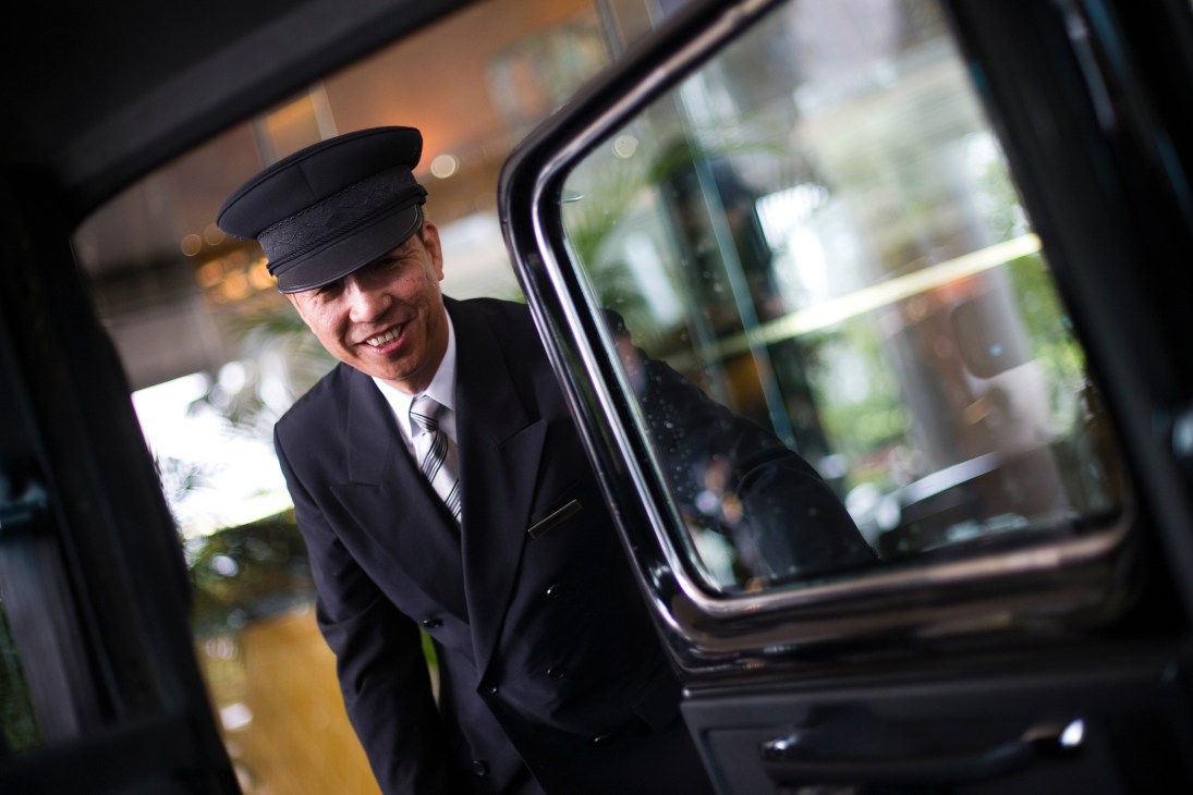 A smiling doorman in uniform and cap opening a car door for a guest.