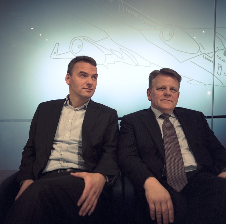 Two men in business suits sit before a wall with aircraft line drawings at IcelandAir offices.