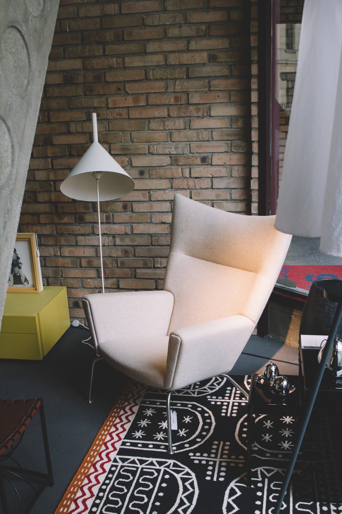 A cream-colored modern armchair with metal legs beside a white floor lamp against a brick wall.