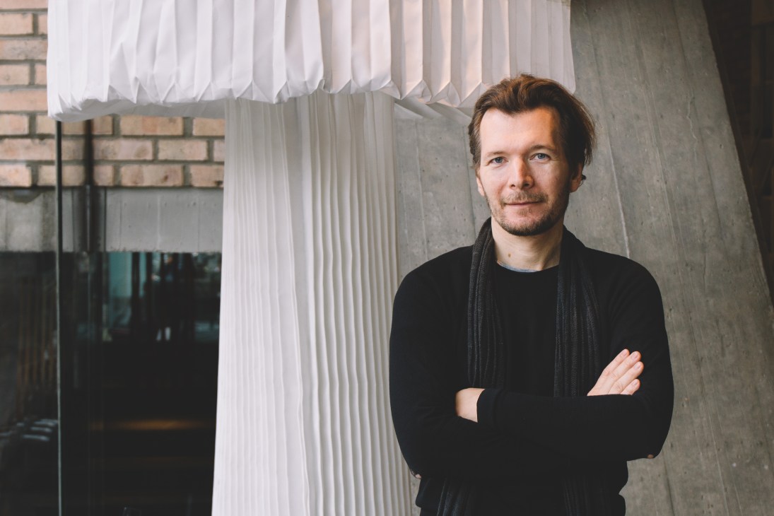 Man in black jacket standing in front of white vertical blinds and concrete wall.